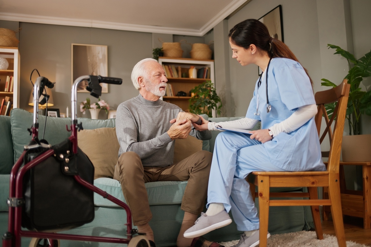 What does a CNA do during a home visit — nurse assistant in blue scrubs holding elderly patient's hand while reviewing care notes