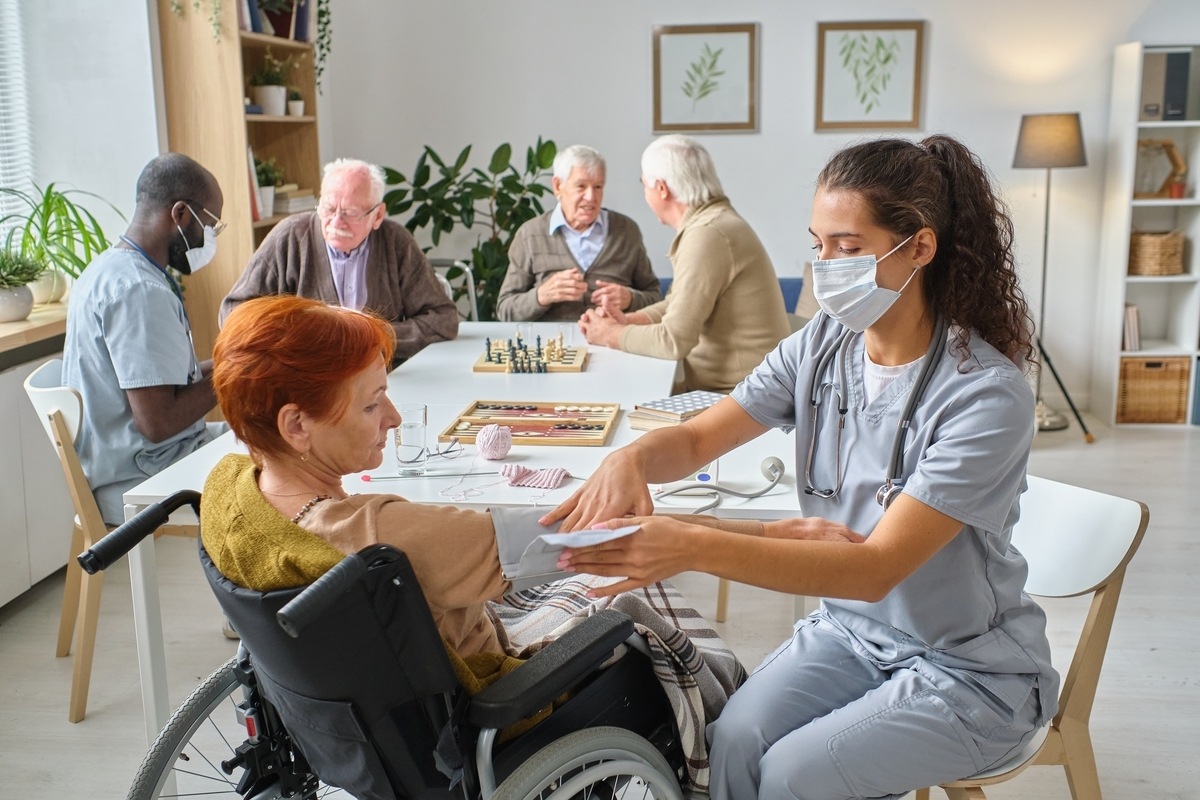 CNA in scrubs assisting an elderly patient in a wheelchair while performing CNA duties in New York at a long-term care facility