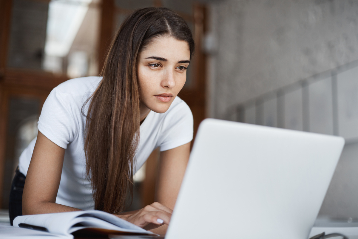 A prospective student researching a medical assistant school in NYC on a laptop with an open book beside her representing the program selection process